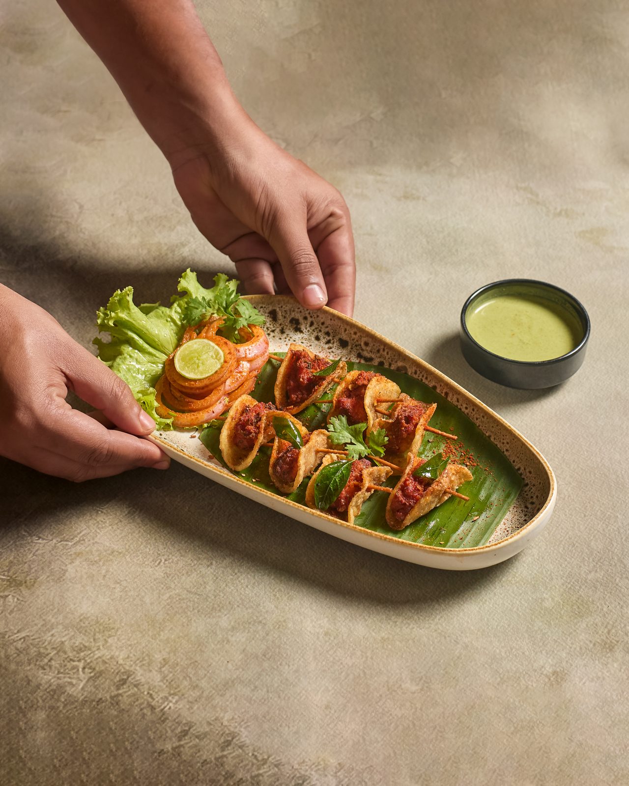A server placing a platter of kebabs on banana leaf with mint chutney